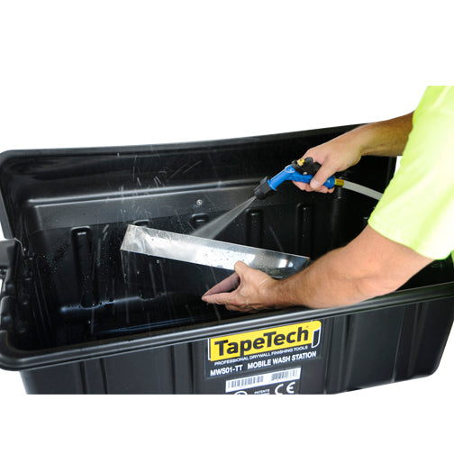 Close-up of a person using a TapeTech water sprayer to clean a stainless steel mud pan inside a black wash basin