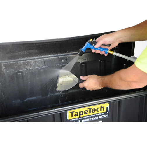 Close-up of a person using a TapeTech water sprayer to clean a stainless steel joint knife inside a black wash basin