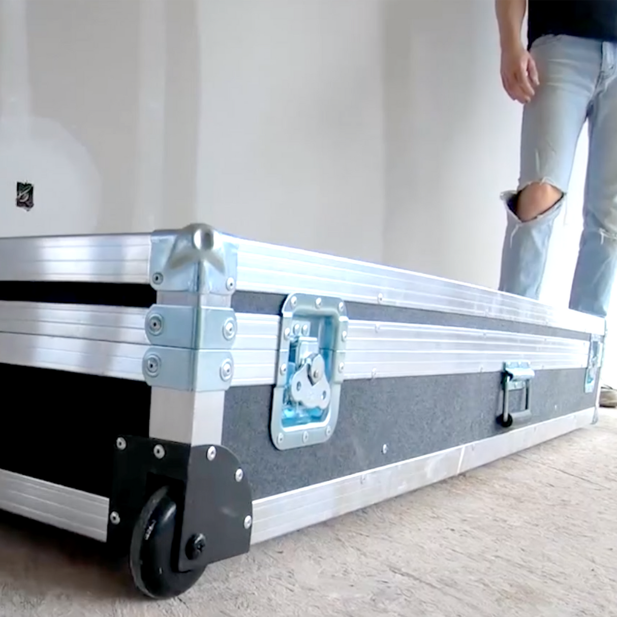 Close-up of a closed auto taping tool case with gunmetal grey trim, latches, and black rolling wheels on the floor at a job site with a worker in the back