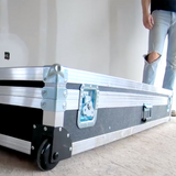Close-up of a closed auto taping tool case with gunmetal grey trim, latches, and black rolling wheels on the floor at a job site with a worker in the back