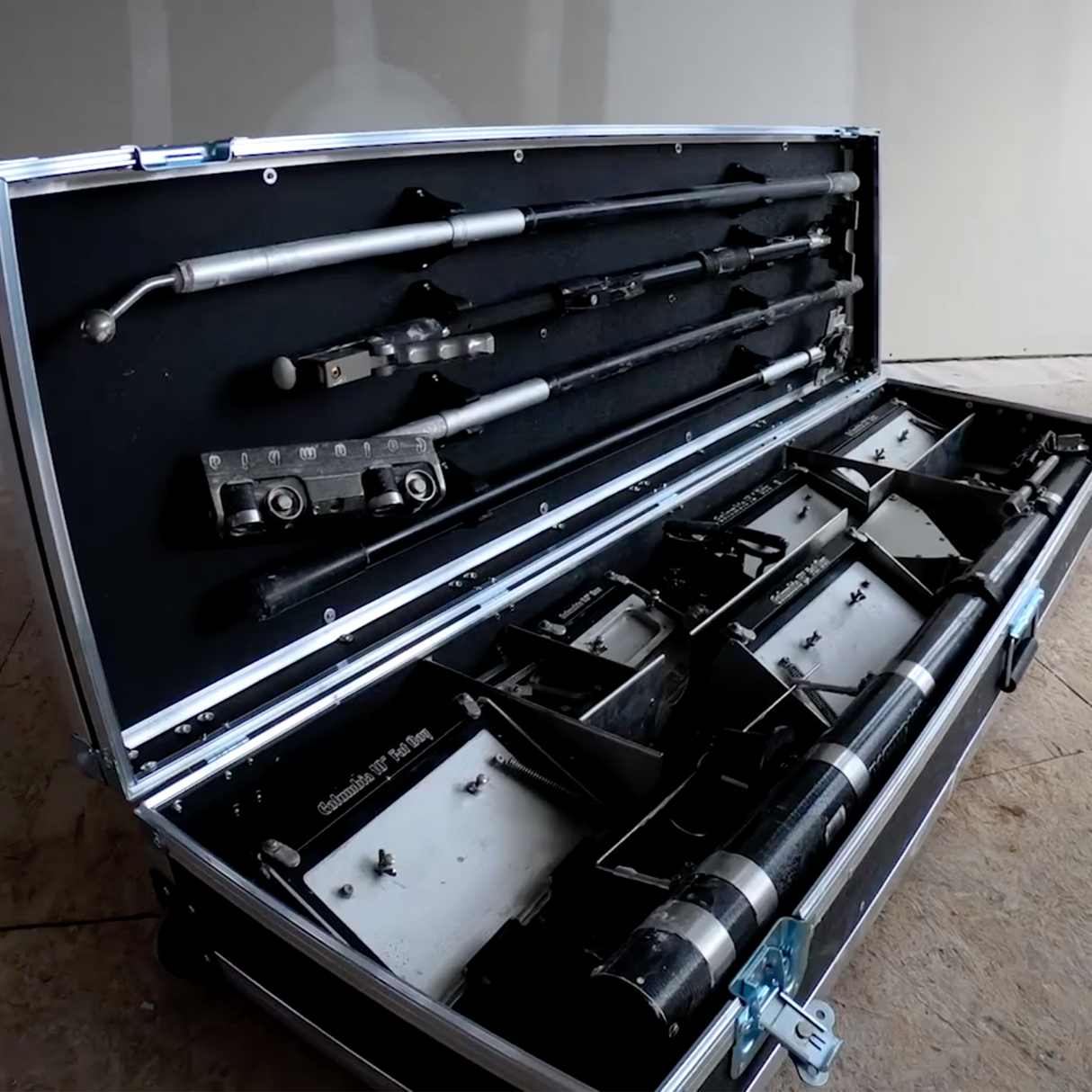Close-up of an open black automatic tool case containing box handles, corner rollers, Fat Boy boxes, and an automatic taper on the floor at a job site